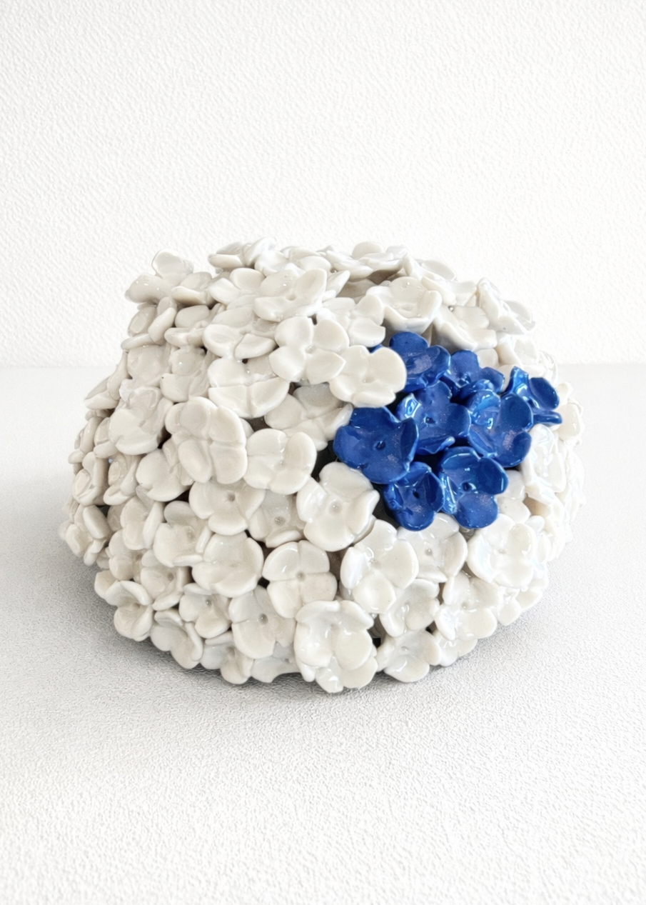 sculptural porcelain wall piece composed of layered white ceramic flowers with subtle shadows, accented by a few deep blue glazed flowers, set against a clean white wall.