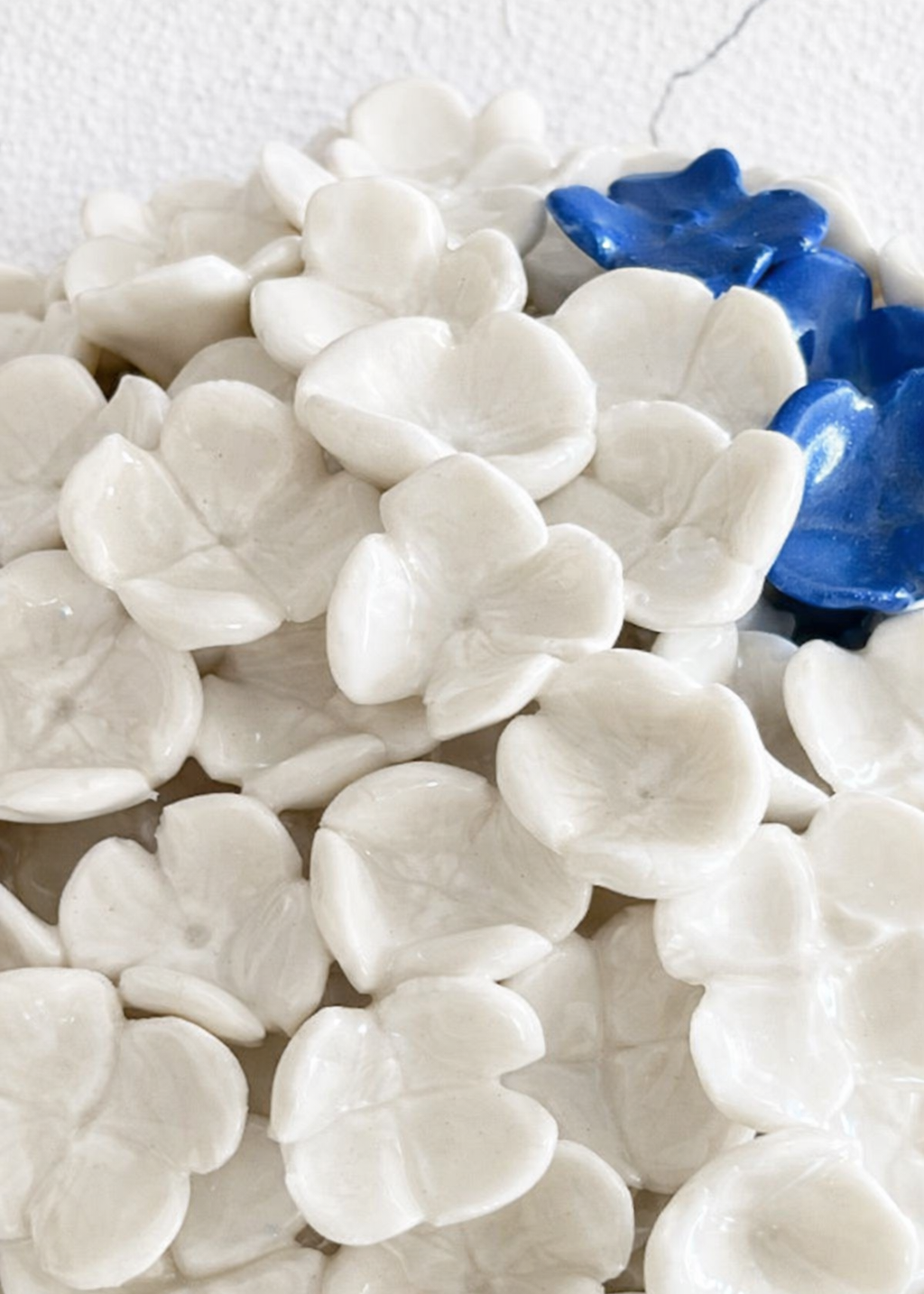 sculptural porcelain wall piece composed of layered white ceramic flowers with subtle shadows, accented by a few deep blue glazed flowers, set against a clean white wall.